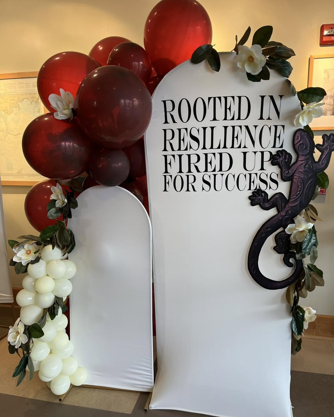 Deep red balloon clusters with ivory florals beside a 'Rooted in Resilience' branded backdrop